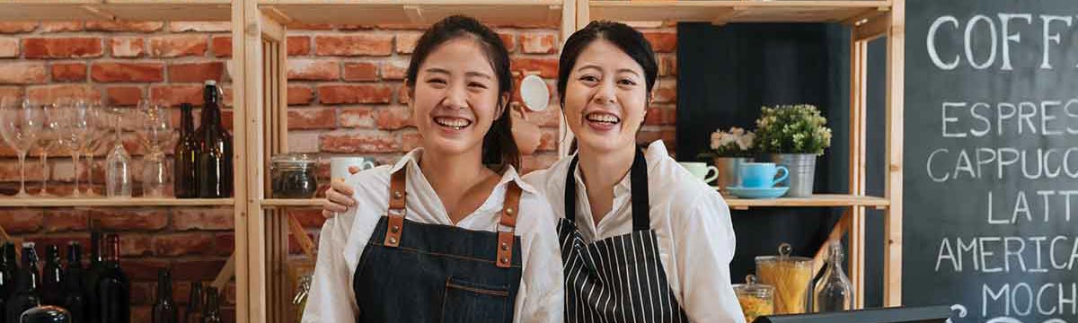 Two successful young baristas women standing in bar counter in cafe in aprons smiling confidently to camera in coffee shop.