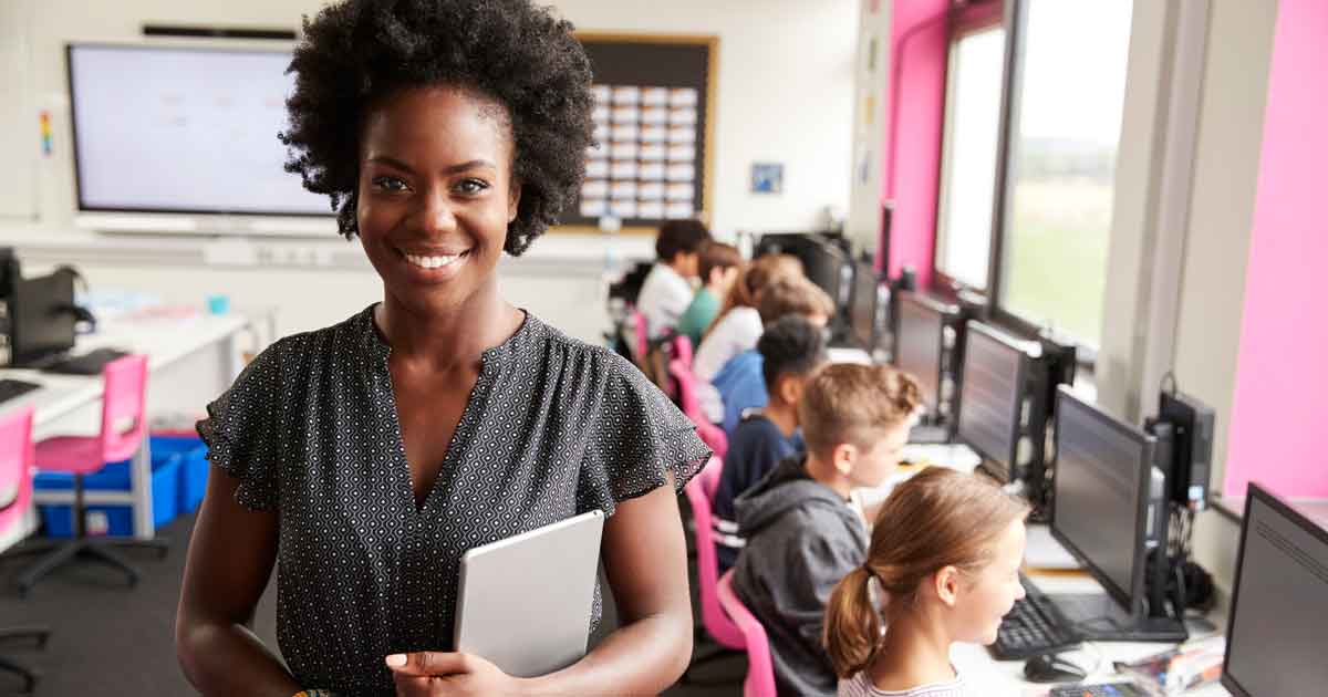 Portrait Of Female Teacher Holding Digital Tablet Teaching Line Of High School Students Sitting By Screens In Computer Class