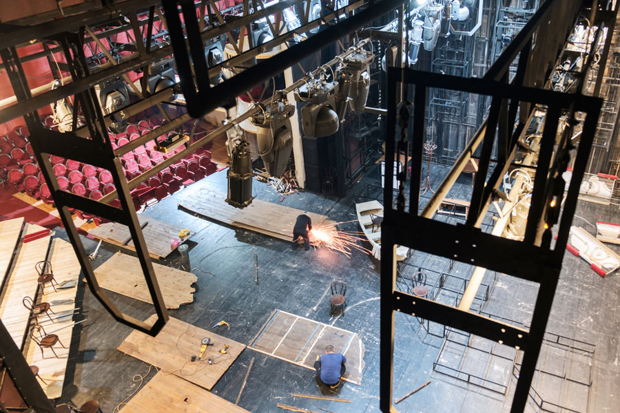 Elevated view of a theater stage with two people working—one welding and one handling wooden planks—amid tools, lighting rigs, and red audience seats.