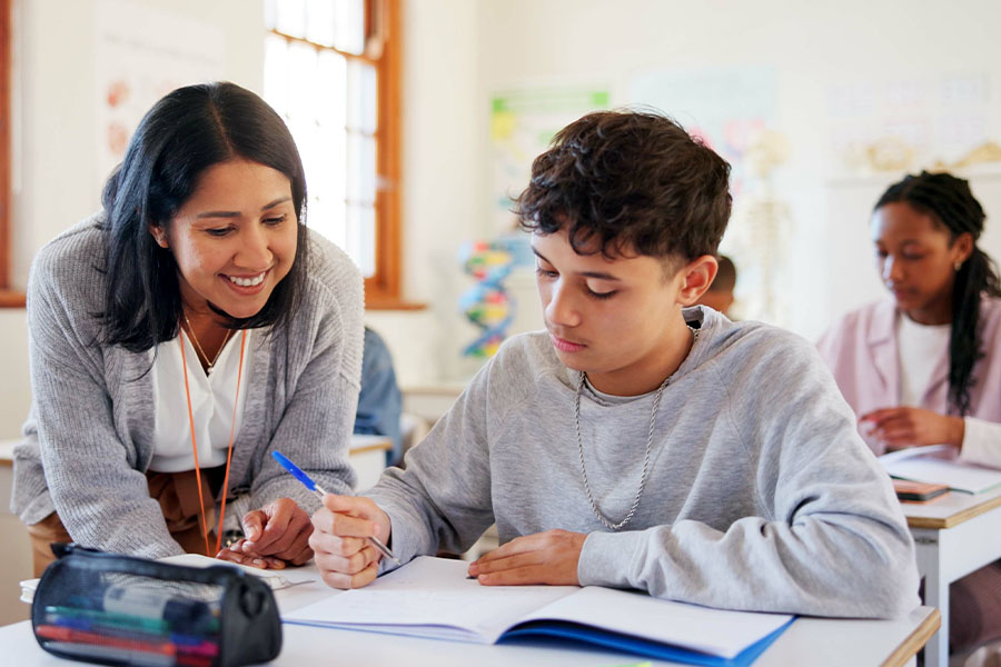 eacher assisting a student at a desk with an open notebook in a classroom; another student is seated in the background.