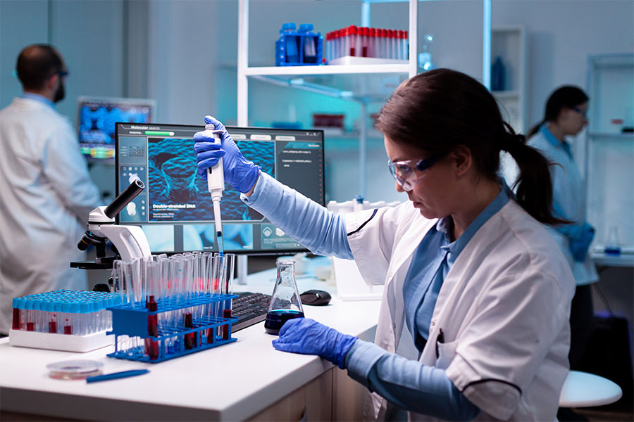 Scientist in white lab coat and blue gloves uses a pipette with test tubes; two others work in the background with lab equipment.