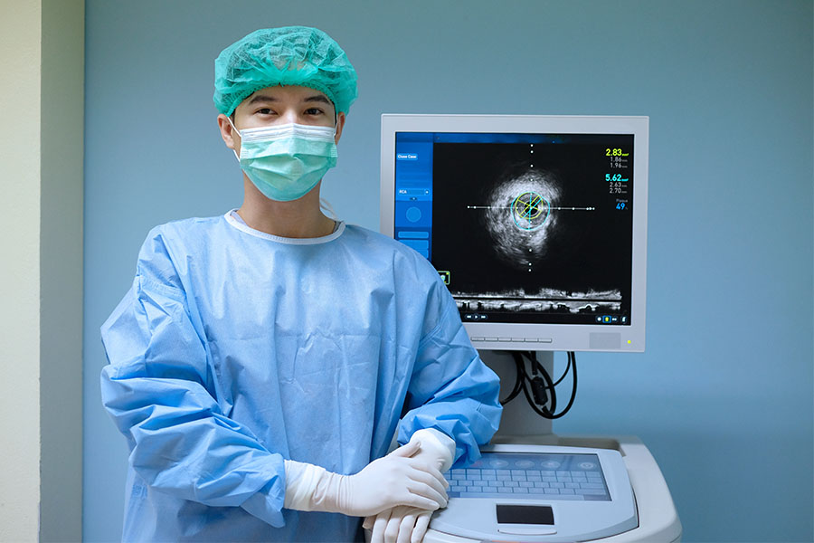 Medical professional in blue gown, white gloves, and green hair cover stands by an imaging machine showing an ultrasound image.