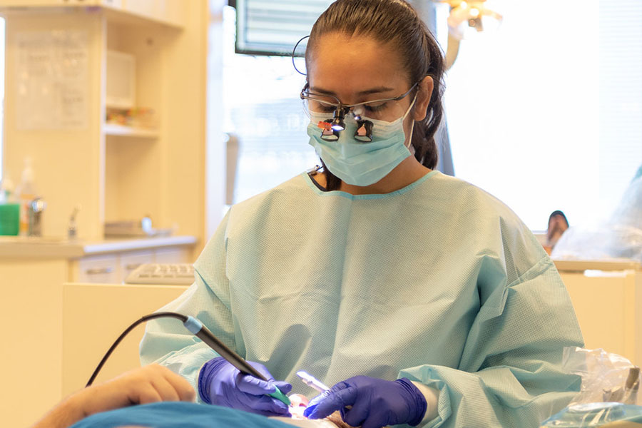 Dentist in blue gown and purple gloves performs a dental procedure; background includes dental office equipment and a window with blinds.